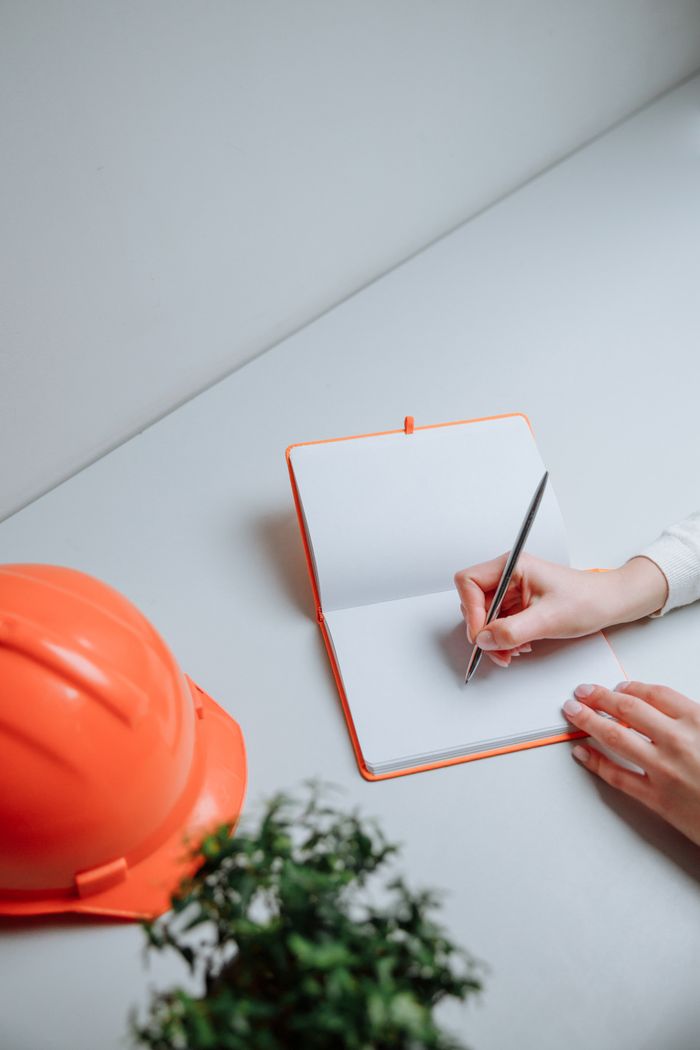 A person writes in a notebook beside an orange hardhat on a white desk.